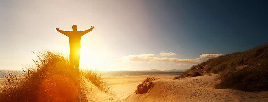 a man standing on a sand dune overlooking the ocean with arms outstretched to God's glory at sunrise 