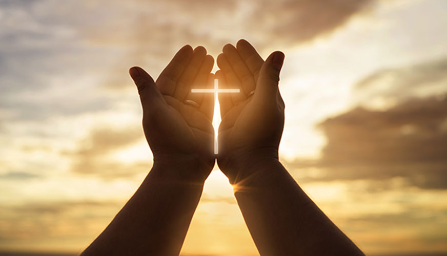 hands held up in prayer in front of the son with a cross of light cupped inside