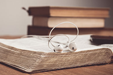 a Bible in the foreground with a pair of white earbuds on top with a stack of books in the background
