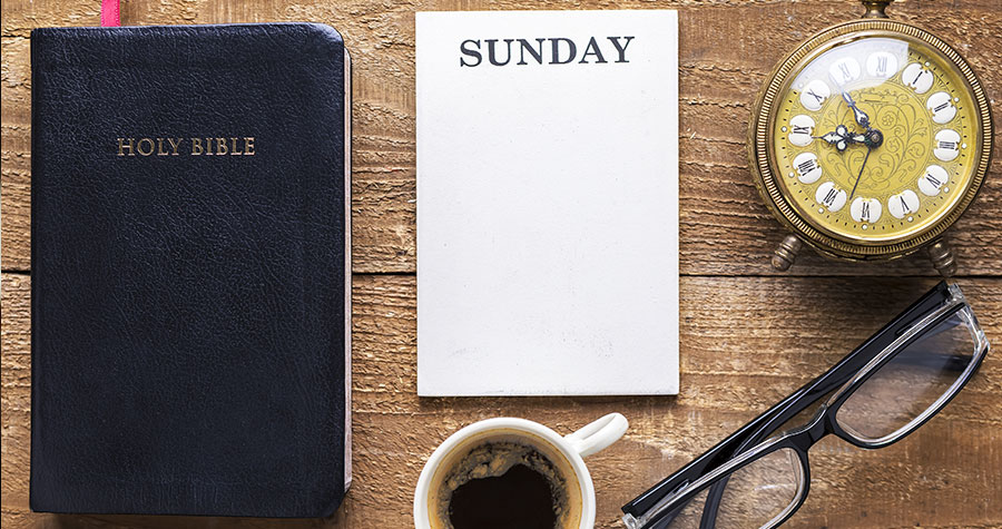 a wooden table with a Bible, a notepad with the word Sunday, an antique pocketwatch, a cup of coffee, and a pair of reading glasses