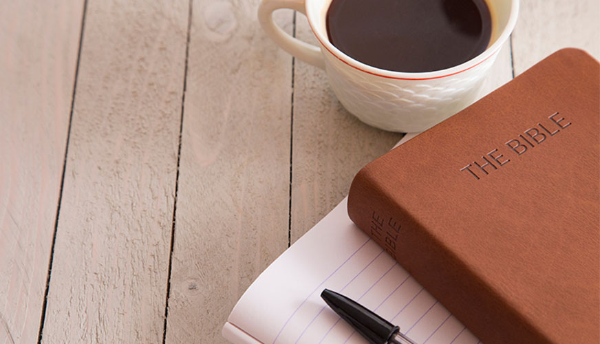 A cup of coffee, a tan leather Bible, a lined notebook, and a pen on top of a light wood plank table