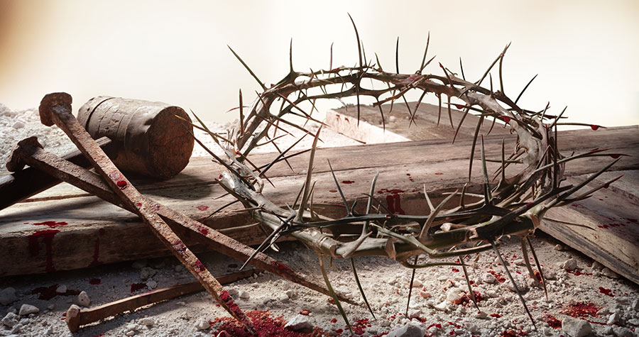 a rough hewn cross held together with thick rop laying on a sandy terrain with a crown of thorns, old bloody rusted nails, and wooden mallet resting on top