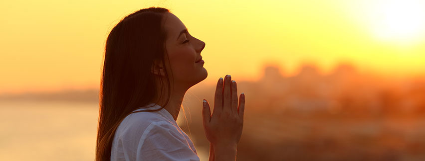 a woman with long straight dark hair praying with eyes closed smiling with the sun rising in the background