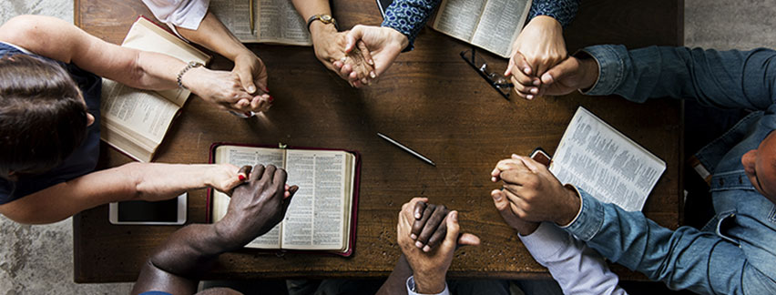 a group of diverse people sitting around a wooden table with open Bibles hands clasped together in prayer